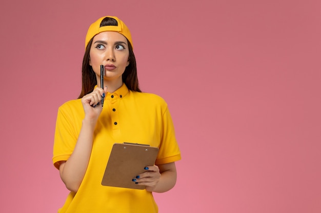 Mensajero femenino de vista frontal en uniforme amarillo y capa sosteniendo bloc de notas y bolígrafo pensando en trabajo de entrega uniforme de servicio de empresa de pared rosa claro