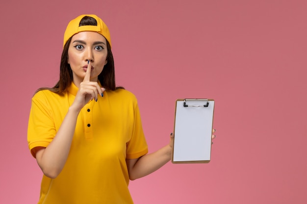 Foto gratuita mensajero femenino de vista frontal en uniforme amarillo y capa sosteniendo el bloc de notas en la entrega uniforme del servicio de la empresa de escritorio rosa