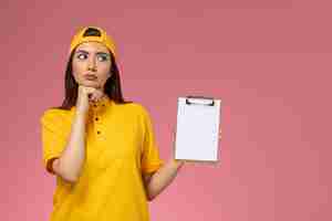 Foto gratuita mensajero femenino de vista frontal en uniforme amarillo y capa sosteniendo el bloc de notas y pensando en la entrega uniforme del servicio de empresa de pared rosa