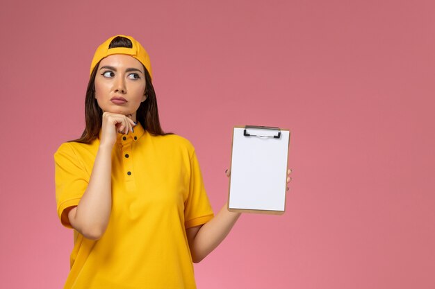 Mensajero femenino de vista frontal en uniforme amarillo y capa sosteniendo el bloc de notas y pensando en la entrega uniforme del servicio de empresa de pared rosa