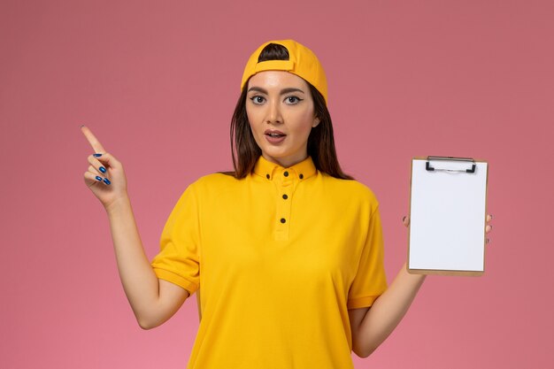 Mensajero femenino de vista frontal en uniforme amarillo y capa sosteniendo el bloc de notas en el trabajo de entrega uniforme de servicio de empresa de pared rosa