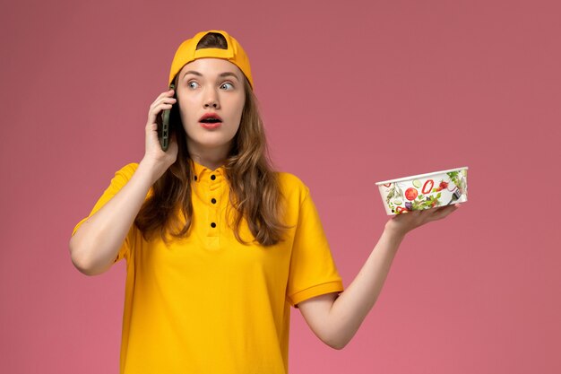 Mensajero femenino de vista frontal en uniforme amarillo y capa sosteniendo cuenco de entrega hablando por teléfono en uniforme de entrega de trabajador de servicio de empresa de pared rosa