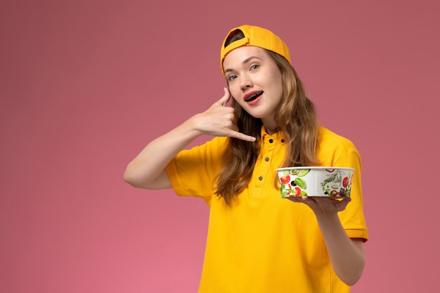 Mensajero femenino de vista frontal en uniforme amarillo y capa sosteniendo el cuenco de entrega en la pared rosa servicio de entrega uniforme de trabajo chica