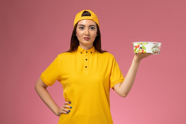 Mensajero femenino de vista frontal en uniforme amarillo y capa sosteniendo un cuenco de entrega redondo y posando en la entrega uniforme del servicio de pared rosa