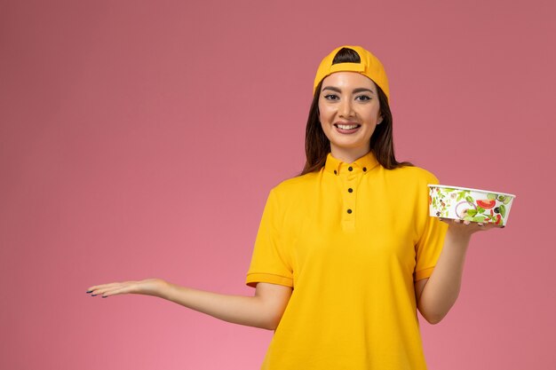 Mensajero femenino de vista frontal en uniforme amarillo y capa sosteniendo un cuenco de entrega redondo y sonriendo en la entrega uniforme del servicio de pared rosa