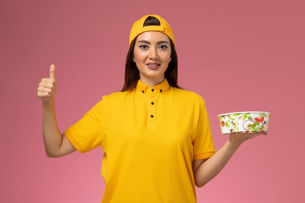 Mensajero femenino de vista frontal en uniforme amarillo y capa sosteniendo cuenco de entrega redondo en el trabajo de entrega uniforme de servicio de pared rosa