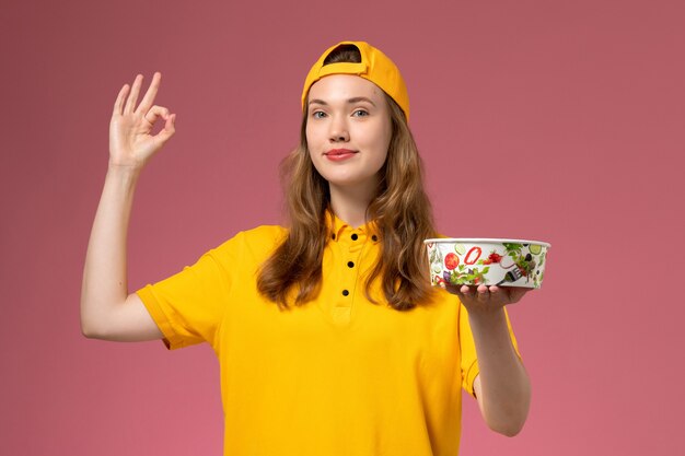 Mensajero femenino de vista frontal en uniforme amarillo y capa sosteniendo el cuenco de entrega en el trabajador de trabajo de empresa uniforme de entrega de servicio de pared rosa