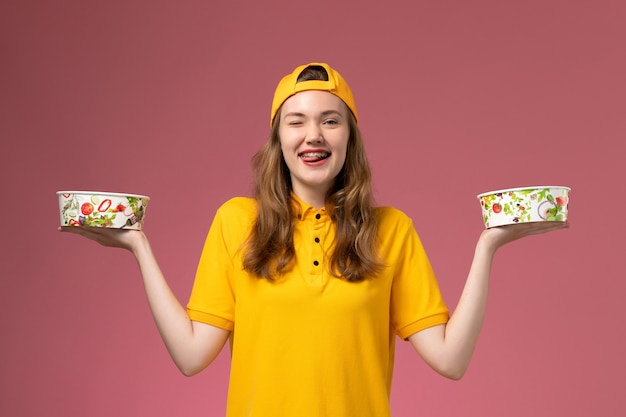 Foto gratuita mensajero femenino de vista frontal en uniforme amarillo y capa sosteniendo cuencos de entrega en la pared rosa trabajador uniforme de trabajo de entrega de servicio