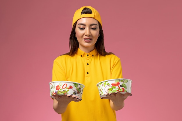 Foto gratuita mensajero femenino de vista frontal en uniforme amarillo y capa sosteniendo cuencos de entrega redondos en trabajo de entrega uniforme de servicio de pared rosa