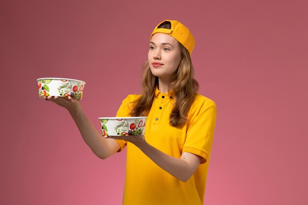 Mensajero femenino de vista frontal en uniforme amarillo y capa sosteniendo cuencos de entrega en servicio de pared rosa uniforme de entrega de trabajo niña trabajadora