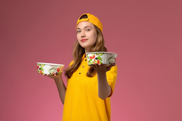 Foto gratuita mensajero femenino de vista frontal en uniforme amarillo y capa sosteniendo cuencos de entrega en el trabajo de entrega de trabajo de servicio de pared rosa