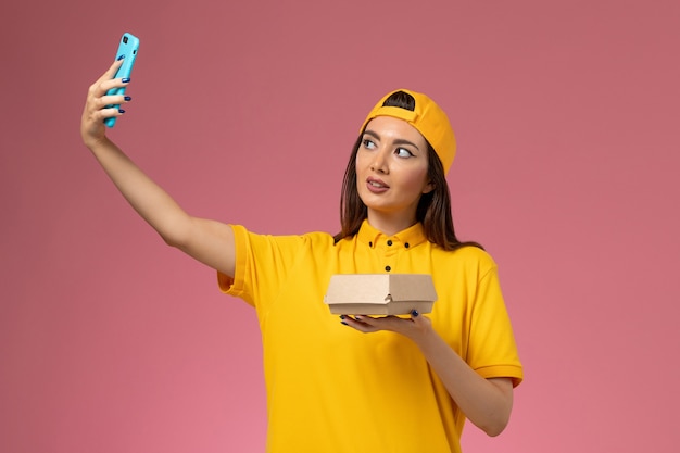 Mensajero femenino de vista frontal en uniforme amarillo y capa sosteniendo el paquete de alimentos y tomando fotos en la entrega uniforme del servicio de la empresa de pared rosa claro