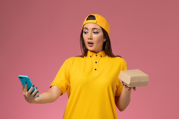 Mensajero femenino de vista frontal en uniforme amarillo y capa sosteniendo el paquete de alimentos y usando un teléfono en el trabajador de entrega uniforme del servicio de la empresa de pared rosa claro