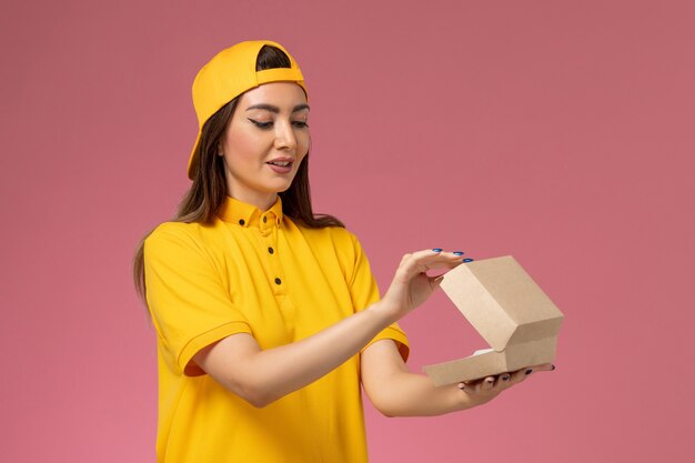 Mensajero femenino de vista frontal con uniforme amarillo y capa sosteniendo un pequeño paquete de comida de entrega y abriéndolo en la empresa de entrega de servicios uniformes de pared rosa