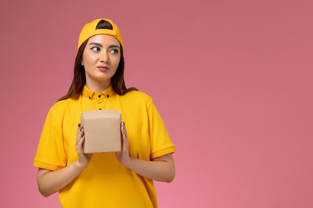 Mensajero femenino de vista frontal con uniforme amarillo y capa sosteniendo un pequeño paquete de comida de entrega en una empresa de entrega de servicio de trabajo uniforme de pared rosa claro