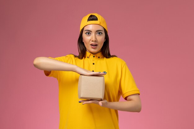 Mensajero femenino de vista frontal en uniforme amarillo y capa sosteniendo un pequeño paquete de comida de entrega en la empresa de trabajo de chica de entrega de servicio uniforme de pared rosa claro