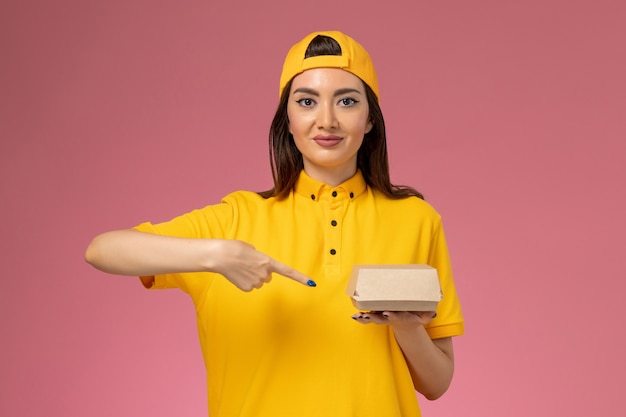 Mensajero femenino de vista frontal en uniforme amarillo y capa sosteniendo un pequeño paquete de comida de entrega en la pared rosa uniforme trabajador de trabajo de la empresa de entrega de servicios