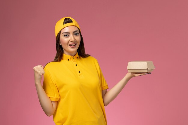 Mensajero femenino de vista frontal en uniforme amarillo y capa sosteniendo un pequeño paquete de comida de entrega en la pared rosa uniforme trabajo de trabajador de entrega de servicio