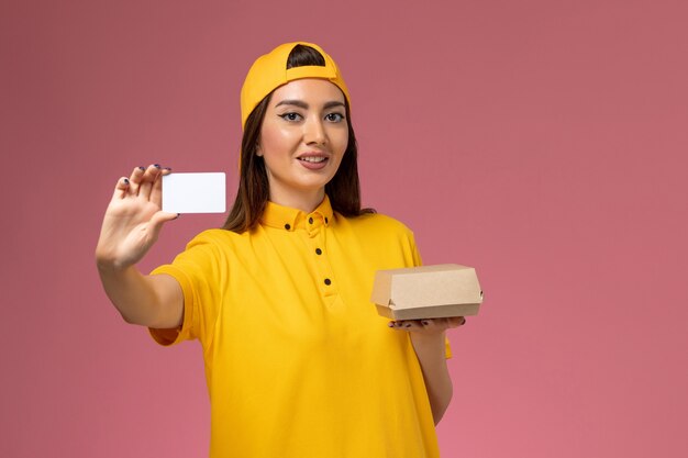 Mensajero femenino de vista frontal con uniforme amarillo y capa sosteniendo un pequeño paquete de comida de entrega y una tarjeta en la empresa de entrega uniforme de trabajo de trabajo de servicio de pared rosa claro