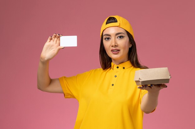 Mensajero femenino de vista frontal en uniforme amarillo y capa sosteniendo un pequeño paquete de comida de entrega y una tarjeta en la empresa de trabajo de entrega uniforme de servicio de pared rosa claro