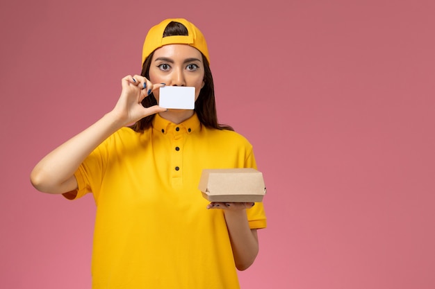 Mensajero femenino de vista frontal con uniforme amarillo y capa sosteniendo un pequeño paquete de comida de entrega y una tarjeta en el trabajo de trabajo de empresa de entrega uniforme de servicio de pared rosa claro