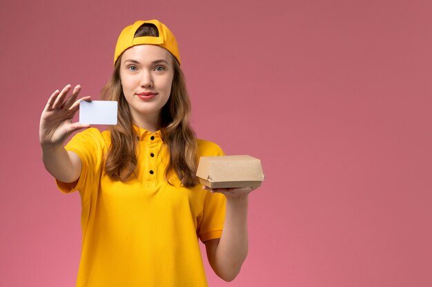 Mensajero femenino de vista frontal con uniforme amarillo y capa sosteniendo un pequeño paquete de comida de entrega y una tarjeta en el uniforme de entrega de trabajo de trabajo de servicio de pared rosa