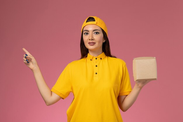 Mensajero femenino de vista frontal con uniforme amarillo y capa sosteniendo un pequeño paquete de comida de entrega en un trabajo de empresa de entrega de servicio uniforme de pared rosa claro