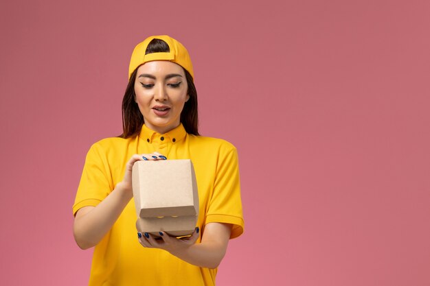 Mensajero femenino de vista frontal en uniforme amarillo y capa sosteniendo un pequeño paquete de comida de entrega en un trabajo de empresa de entrega uniforme de servicio de pared rosa claro