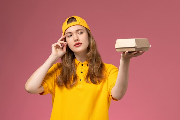 Mensajero femenino de vista frontal en uniforme amarillo y capa sosteniendo un pequeño paquete de comida de entrega en el trabajo uniforme de entrega de servicio de pared rosa