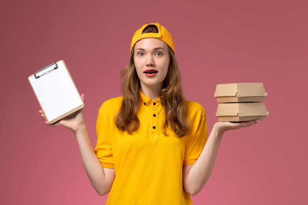 Mensajero femenino de vista frontal en uniforme amarillo y capa sosteniendo pequeños paquetes de comida de entrega con bloc de notas en la pared rosa trabajo de trabajo uniforme de entrega de servicio