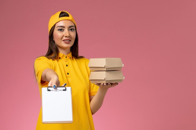 Mensajero femenino de vista frontal en uniforme amarillo y capa sosteniendo pequeños paquetes de comida de entrega con bloc de notas en el trabajo de entrega uniforme de servicio de pared rosa