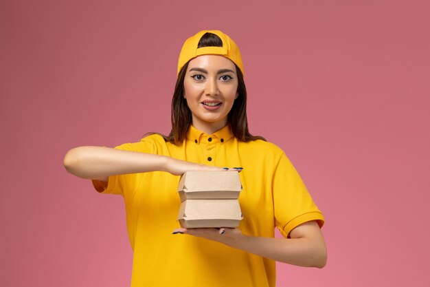 Mensajero femenino de vista frontal con uniforme amarillo y capa sosteniendo pequeños paquetes de comida de entrega y sonriendo en la pared rosa claro servicio de entrega uniforme