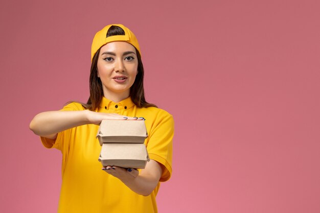 Mensajero femenino de vista frontal en uniforme amarillo y capa sosteniendo pequeños paquetes de comida de entrega en el trabajo de trabajo de entrega uniforme de servicio de pared rosa claro