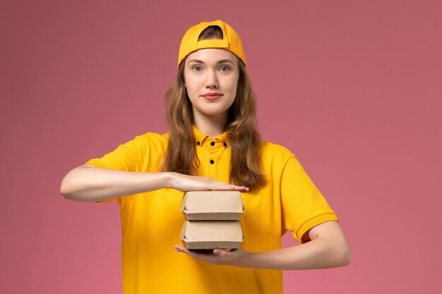 Mensajero femenino de vista frontal en uniforme amarillo y capa sosteniendo pequeños paquetes de comida de entrega en el trabajo de trabajo uniforme de entrega de servicio de pared rosa