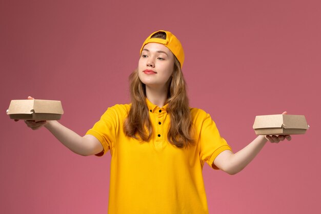 Mensajero femenino de vista frontal con uniforme amarillo y capa sosteniendo pequeños paquetes de comida de entrega en uniforme de entrega de trabajo de servicio de pared rosa claro