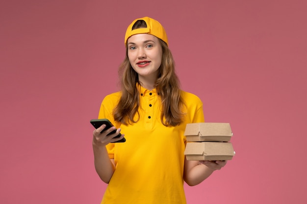 Mensajero femenino de vista frontal en uniforme amarillo y capa sosteniendo teléfono con paquetes de alimentos sonriendo en uniforme de entrega de servicio de empresa de pared rosa