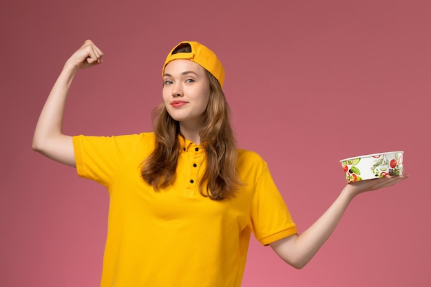 Mensajero femenino de vista frontal en uniforme amarillo y capa con tazón de entrega flexionando en la pared rosa trabajo de empresa uniforme de entrega de servicio