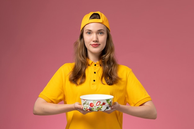 Mensajero femenino de vista frontal en uniforme amarillo y capa con tazón de entrega en la pared rosa trabajo de empresa uniforme de entrega de servicio