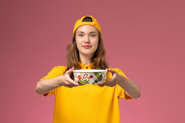 Mensajero femenino de vista frontal en uniforme amarillo y capa con tazón de entrega en la pared rosa trabajo de empresa uniforme de entrega de servicio