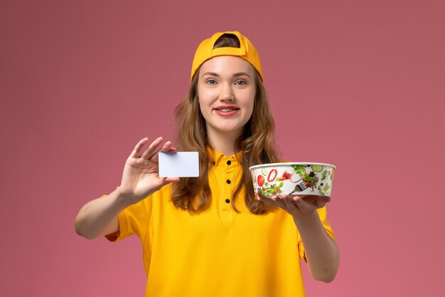 Mensajero femenino de vista frontal en uniforme amarillo y capa con tazón de entrega y tarjeta en el trabajo de empresa uniforme de entrega de servicio de pared rosa