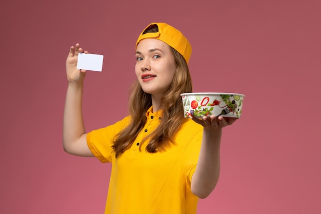 Mensajero femenino de vista frontal en uniforme amarillo y capa con tazón de entrega y tarjeta en el trabajo de trabajador de empresa uniforme de entrega de servicio de pared rosa