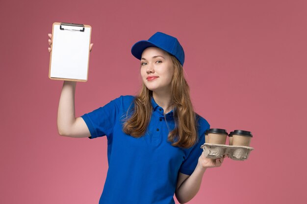 Mensajero femenino de vista frontal en uniforme azul con bloc de notas y tazas de café de entrega con sonrisa en la empresa de uniforme de servicio de escritorio rosa claro