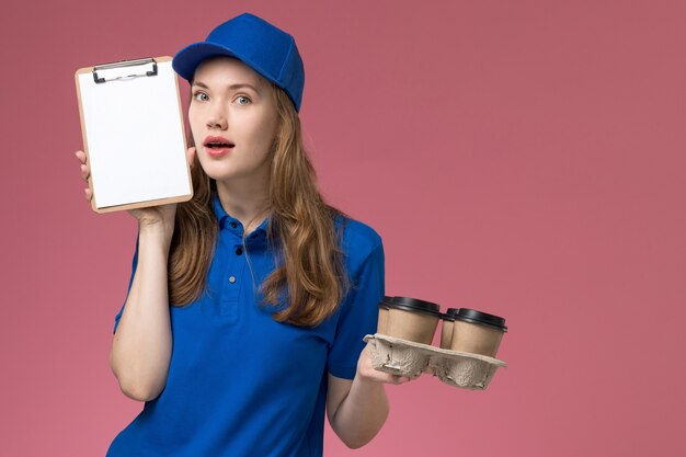 Mensajero femenino de vista frontal en uniforme azul con bloc de notas y tazas de café de entrega en el trabajo de la empresa uniforme de servicio de escritorio de luz rosa