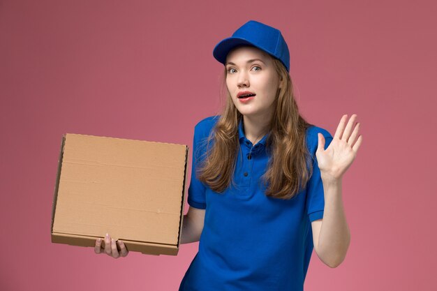 Mensajero femenino de vista frontal en uniforme azul con caja de comida con expresión de sorpresa en la empresa de uniforme de servicio de trabajo de escritorio rosa