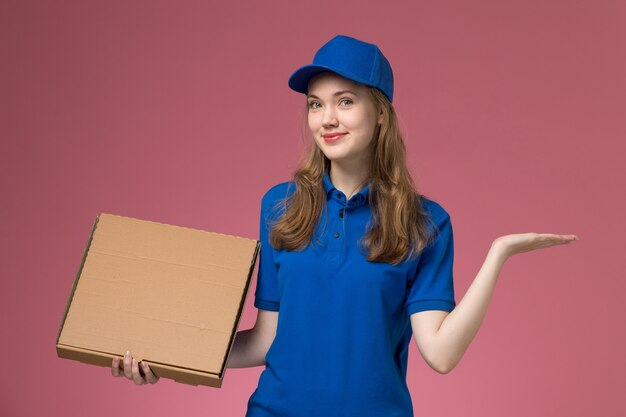 Mensajero femenino de vista frontal en uniforme azul con caja de comida con mano rasied sobre fondo rosa empresa uniforme de servicio de trabajo