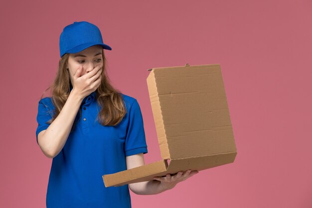 Mensajero femenino de vista frontal en uniforme azul con caja de entrega de alimentos abriéndola con expresión de sorpresa en el trabajo de la empresa uniforme de servicio de trabajador de escritorio rosa