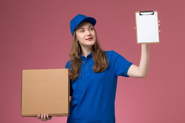 Mensajero femenino de vista frontal en uniforme azul con caja de entrega de alimentos y bloc de notas con una sonrisa en el trabajador de la empresa uniforme de servicio de escritorio rosa