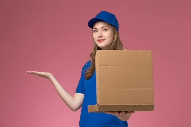 Mensajero femenino de vista frontal en uniforme azul con caja de entrega de alimentos posando con él en la empresa de uniforme de servicio de trabajo de fondo rosa