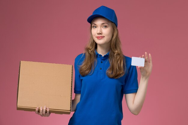 Mensajero femenino de vista frontal en uniforme azul con caja de entrega de alimentos y tarjeta blanca en el trabajo de empresa uniforme de servicio de escritorio rosa