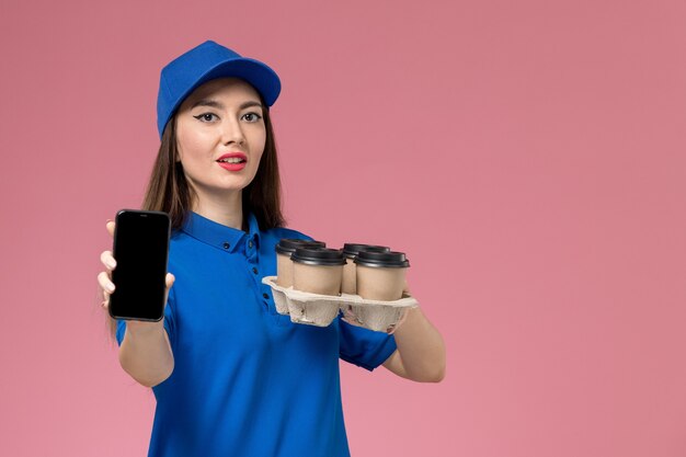 Mensajero femenino de vista frontal en uniforme azul y capa mostrando teléfono y sosteniendo tazas de café en la pared rosa